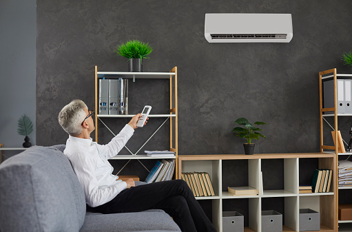 Senior man switches AC in his workplace. Older businessman sitting on couch in his modern office interior turns on electronic wall air conditioner using remote control. Comfort and lifestyle concept