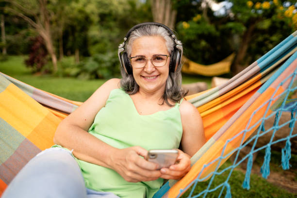 Smiling mature woman sitting in a hammock listening to music on headphones Portrait of a smiling mature woman sitting in a hammock in summer and listening to a smart phone playlist on headphones hammock relaxation women front or back yard stock pictures, royalty-free photos & images