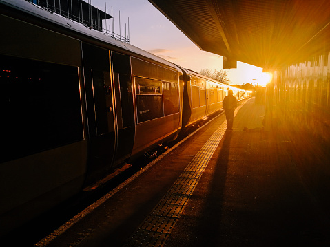 Color image depicting commuters (silhouetted and rendered unrecognizable by the glowing setting sun) walking next to a passenger train on a station platform at sunset.