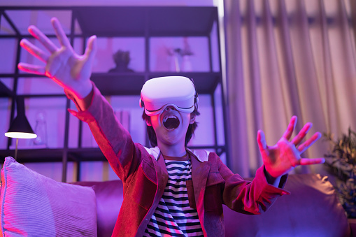 Asian child boy wear VR or virtual reality glasses,headsets standing and playing a video game at living room in front of TV at home in quarantine period technology and innovation concept neon light night lifestyle