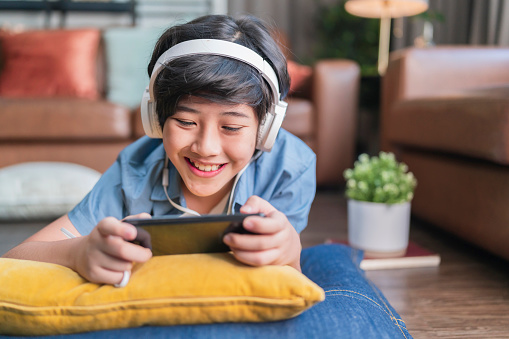 chilling weekend holiday with esport game online,asian child boy wear headphone hand playing smartphone game online with exited and fun laydown on floor at living room at home, son is playing alone with mobile equipment.