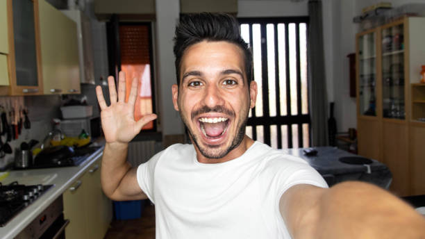 jovem bonito tirando selfie com celular inteligente em casa - cara feliz sorrindo para a câmera - influenciador, blogueiro de vídeo e conceito de namoro online - mobile phone home video camera video photograph - fotografias e filmes do acervo