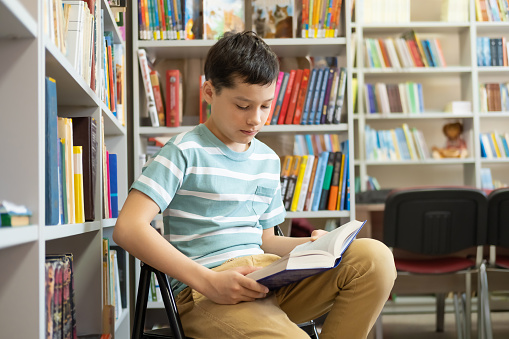 Caucasian teenage schoolboy boy reads book in modern public library. Concept of school and additional education