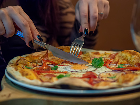Woman's hand holding fork and knife and cutting a delicious thin crust pizza. Close up of pizza plate with fry egg, cheese and vegetables. Dinner with friends in Italian restaurant.