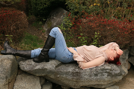 A Caucasian woman lying on a rock in a garden. She is wearing a pink top, torn blue jeans and black boots.
