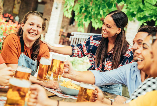 счастливые друзья звенят пивными бокалами в саду паба пивоварни - молодые люди, сидящие за барным столом, едят закуски и пьют алкоголь вмест - parks brewery стоковые фото и изображения