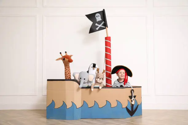 Photo of Cute little boy playing with toys in pirate cardboard ship near white wall indoors