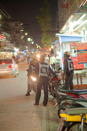 Group of thai police motorcycle patrol is standing in street at night. Men are talking, some are standing, some are sitting on motorcycle. They are in front of a small supermarket in residential district in Bangkok Chatuchak
