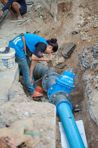 Thai blue worker woman is counter fixing screws of water pipe in construction site in street where new water pipes are being laid out. Scene is in Bangkok Ladprao