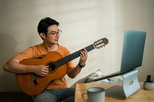 Medium shot Asian male playing guitar with her little daughter Teleconferencing with music with his friend via video call on internet. Man having fun playing classical guitar. Musician man teaching online guitar lesson with his student on laptop computer. Online music class