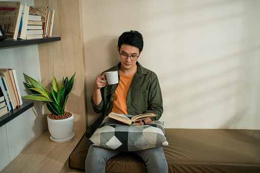 Asian Man Smiling while  reading a book as hobby in the living room at home in the weekend.