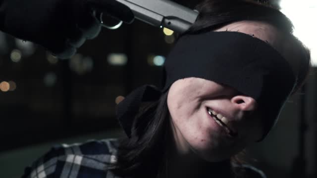 Close up criminal put a gun to the head of a tied young woman blindfolded