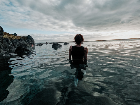 Single Caucasian female waist deep in the warm geothermal lagoon waters near Reykjavik, Iceland. Towering boulders and turquoise waters surround her. Rear view.