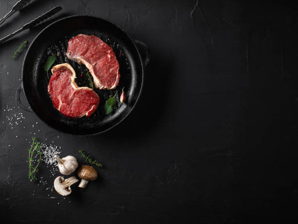 raw steaks with seasonings in an iron grilling pan, black background. fresh meat, herbs, garlic, pepper, salt, champignon, tongs. close-up. top view - top sirloin imagens e fotografias de stock