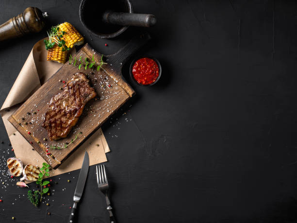 Fried steak with herbs and spices on wooden board, parchment, fork, knife, garlic, corn, sprouts, pepper, salt, sauce. Black background. Top view stock photo