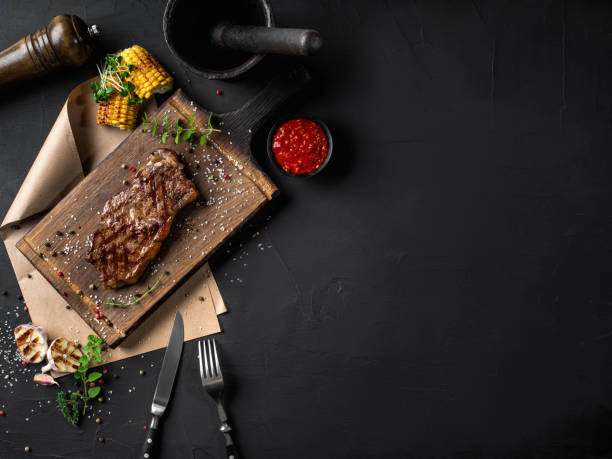 Fried steak with herbs and spices on wooden board, parchment, fork, knife, garlic, corn, sprouts, pepper, salt, sauce. Black background. Top view Fried steak with herbs and spices on a wooden board, parchment, steel fork and knife, grilled garlic and corn, sprouts, spice chopper, sauce in a small dark bowl, mortar with pestle on a black background. Cooking, restaurant concept. Close-up shot. Top view. Dry aged rosemary seedlings stock pictures, royalty-free photos & images