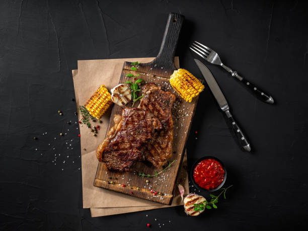 istock Fried steaks with herbs and spices on wooden board, parchment, grilled garlic, corn, sauce, fork and knife on black background. Close-up. Top view 1393219336
