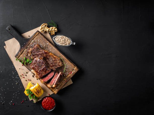 Fried steaks with herbs and spices on wooden board, parchment, grilled garlic, corn, two sauces on black background. Close-up. Top view stock photo