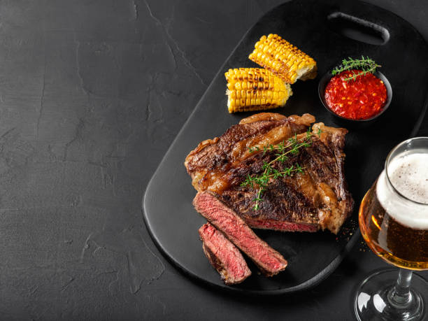 Sliced, fried, spiced juicy steaks with herbs on dark board, grilled corn, red sauce and glass of beer on black background. Close-up shot. Side view stock photo