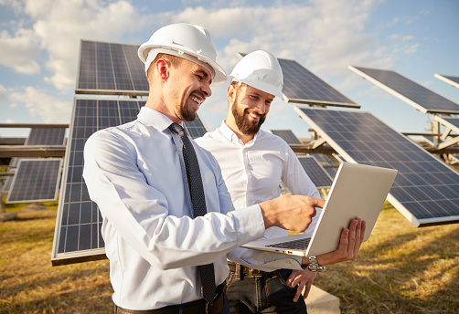 Positive male technicians in formal wear and hardhats discussing configuration of solar panels while standing close and using netbook together