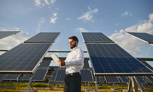 Low angle of bearded male investor analyzing data on tablet while standing near photovoltaic panels on sunny day on solar farm