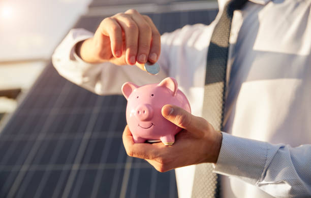 Man putting money into piggy bank near solar panel stock photo