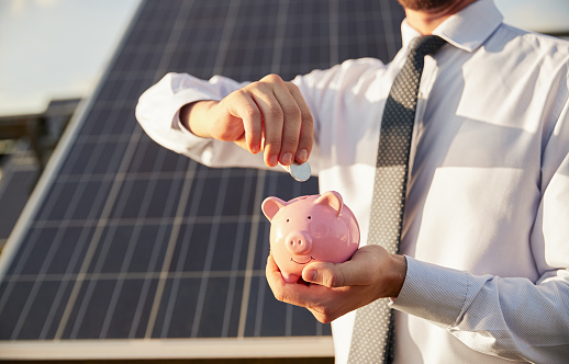 Unrecognizable male manager putting coin into piggy bank while saving money near photovoltaic panel on solar farm