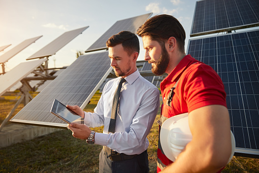 Concentrated young businessman in formal outfit showing project details on tablet to male technician standing near photovoltaic panels on solar power station