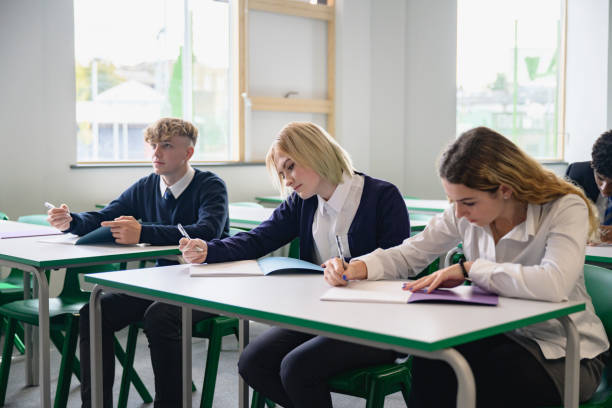 Students taking educational exam in secondary classroom Three-quarter front view of male and female teenagers in uniforms sitting at desks and concentrating while writing in composition booklets. teenagers only teenager multi ethnic group student stock pictures, royalty-free photos & images