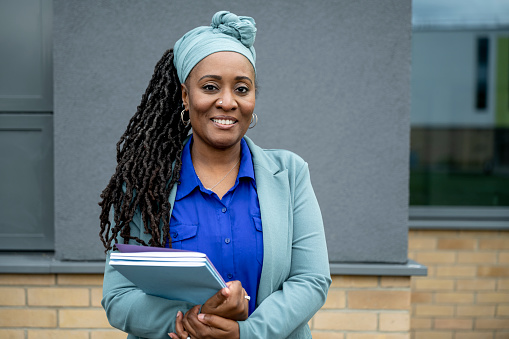 Waist-up front view of mature Black woman with locs hairstyle in business attire and headscarf standing outdoors on school campus smiling at camera.