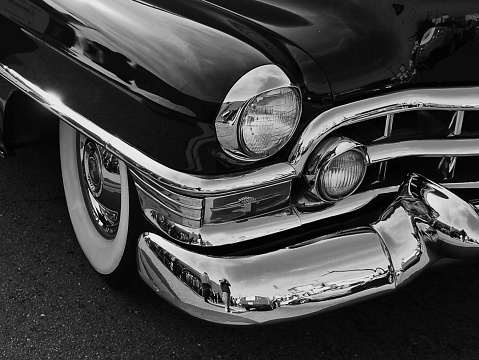 Black and white photograph.  Car headlight on black old car with chrome bumper.  White wall tires.  Rounded fenders and rounded bumpers.