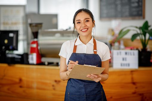 Opening a small business, AHappy Asian woman in an apron standing near a bar counter coffee shop, Small business owner, restaurant, barista, cafe, Online, SME, entrepreneur, and seller concept