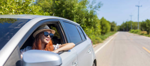 Woman with ginger red hair wearing sunglasses driving car in solo weekend getaway vacation trip looking out the window in summer with green nature in countryside road for freedom and adventure mujer con cabello rojo jengibre con gafas de sol conduciendo coche en solitario escapada de fin de semana viaje de vacaciones mirando por la ventana en verano con exuberante naturaleza verde en el camino del campo para la libertad y la aventura