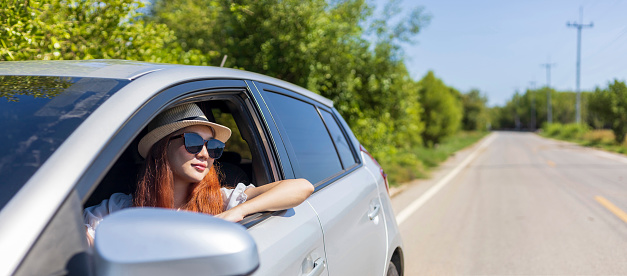 Woman with ginger red hair wearing sunglasses driving car in solo weekend getaway vacation trip looking out the window in summer with green nature in countryside road for freedom and adventure