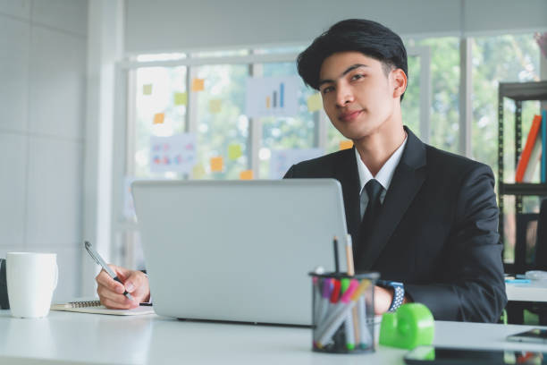 istock Asian young business man is writing while working with laptop in office, looking at camera. 1393253625