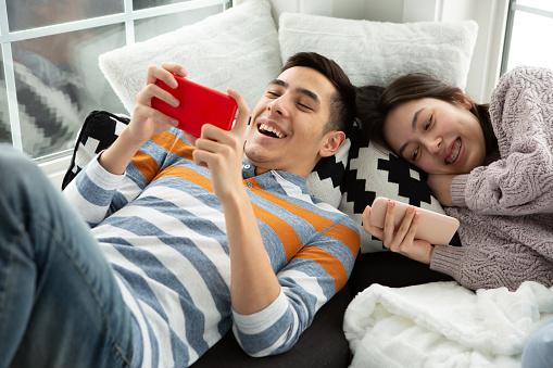 Smiling mixed race brother and sister relaxing and using smart phone laying in living room nook corner.