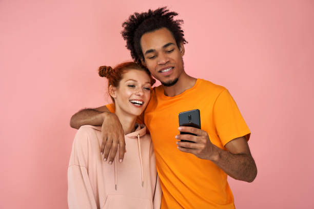 Young mixed race couple embrace each other, look joyfully at mobile phone, glad to view photos Positive young mixed race couple embrace each other, look joyfully at mobile phone, glad to view common photos, satisfied with good wireless connection, against pink background. Relationship concept. unusual couple stock pictures, royalty-free photos & images