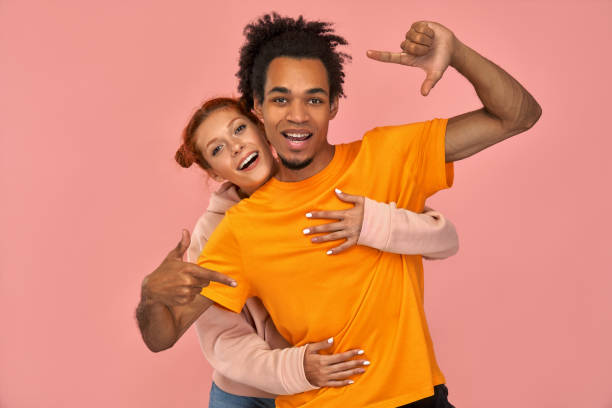 istock Happy young dark skinned man with cheerful expression, makes hand frame, tries to find right perspective, searches inspiration or right angle, his ginger girlfriend hugging behind, pink background 1393261109