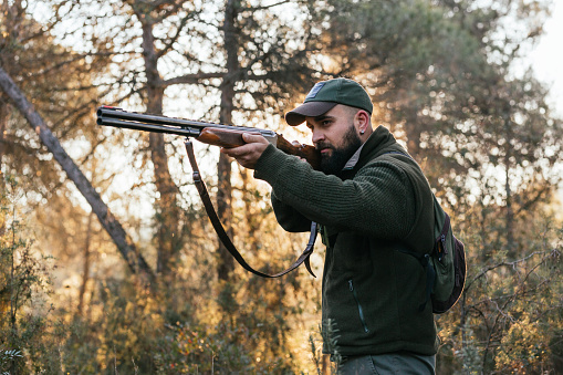 Male hunter with backpack aiming rifle and attentively looking at distance while hunting in forest in countryside