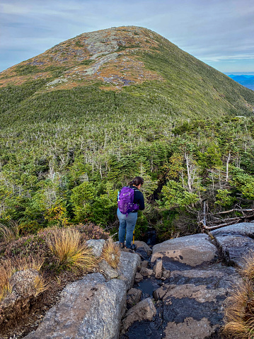 A female hiker looking to complete her 46 high peak challenge in Adirondack park New York looks down a rocky wet cliff on Iroquois peak towards Algonquin peak in the distance