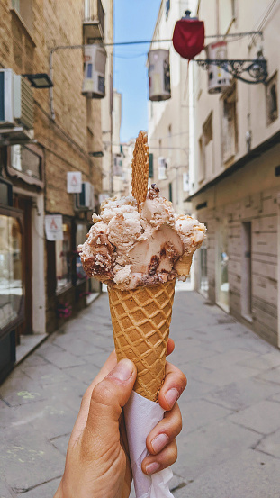 Hand holding a vegan cream cream cone with biscuit waffle in a street in Alghero, Italy on a sunny day