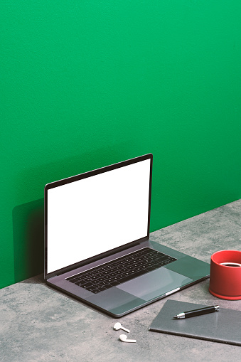 Laptop mockup, template on concrete table top with green background. Red mug, earphones, notepad and pencil on the table.