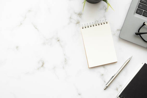 istock Blank small notebook with pen are on top of white marble office desk table with laptop computer and office supplies. Top view with copy space, flat lay. 1393355794