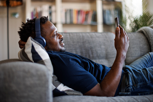 Smiling man with headphones lying on his sofa in the living room at home and using his smart phone