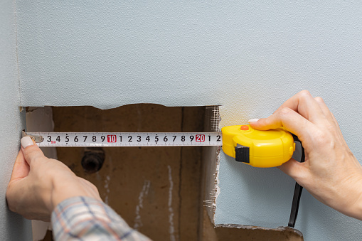a woman measures a hole in the wall with a tape measure