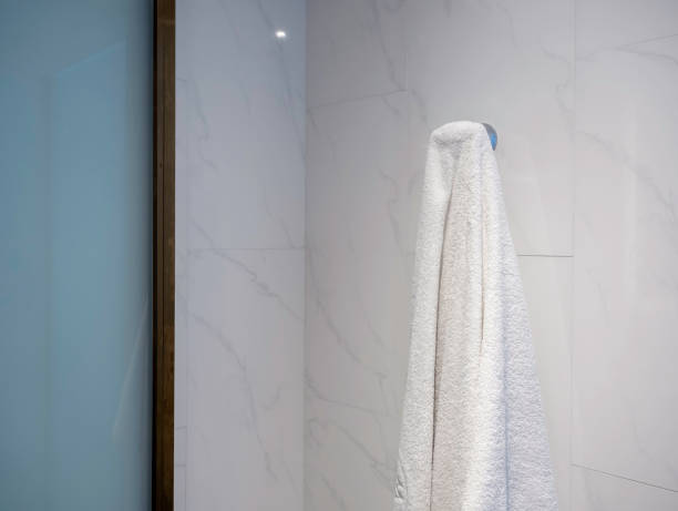 istock Close-up white clean towel hanging on a hook, hanger on white marble wall background in a luxury hotel room near the glass door of shower room. 1393368462
