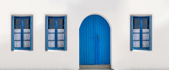 Wooden door closed and three glass windows with open shutters blue color on white wall background. Cyclades island house front view, Greek traditional architecture.