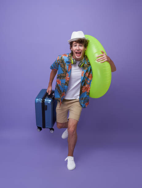 Excited hurry young man tourist with luggage and rubber ring running to holiday isolated on purple studio background. stock photo