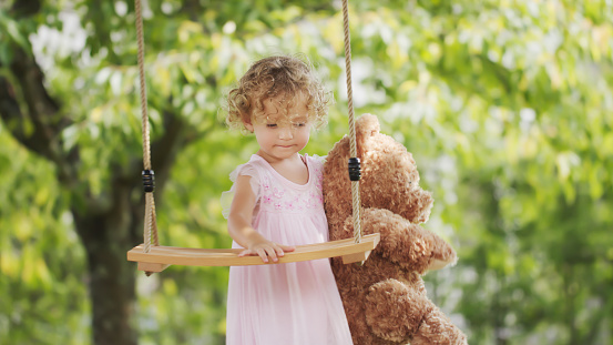 Spring and happy summer time. Joyful smiling little girl playing with teddy bear swinging on the swing. Child with blue eyes and curly blond hair in the green garden at home, concept of healthy growth