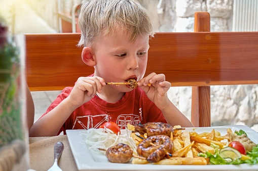 Toddler eats meat with fries and vegetables on restaurant terrace in fresh air. Blond boy enjoys dinner on holiday in Croatian city Omis closeup
